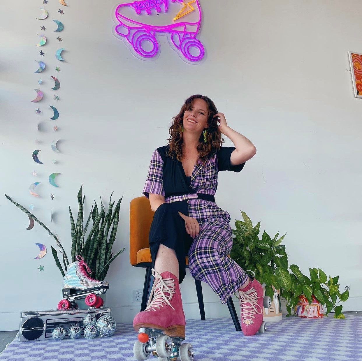 Stacey Skates wears a black and purple plaid jumpsuit with pink roller skates. She sits on a chair surrounded by plants and the wall behind features a neon roller skate light at the RollerFit Shop.