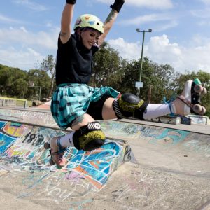 Zoe wears a black tshirt, blue skirt, white helmet, leopard print protective gear and pink roller skates. She is doing a big fun forward kick air at the skatepark.