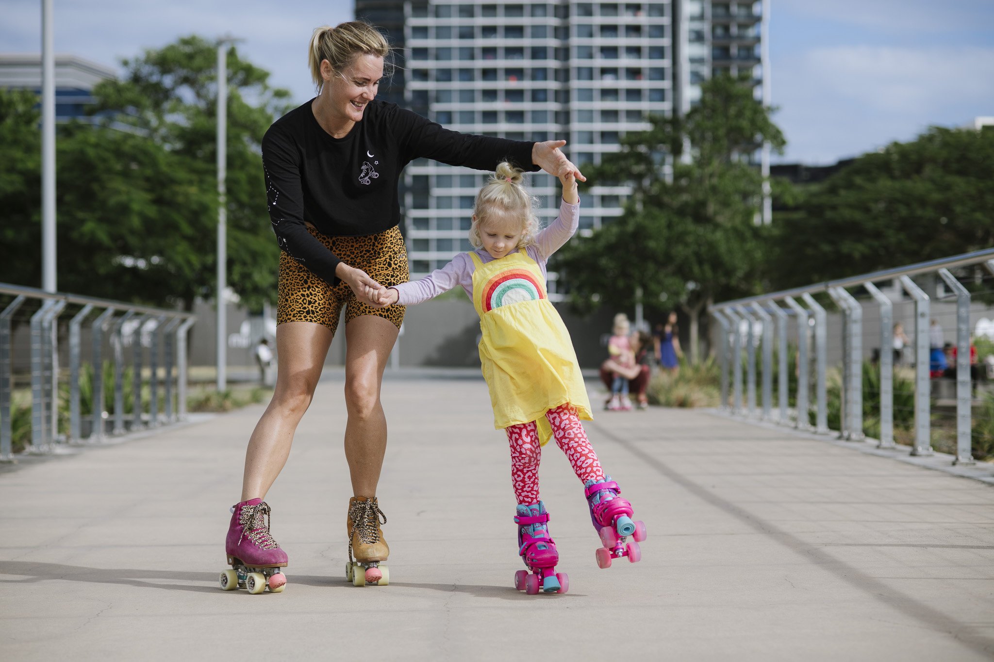 Nicola from RollerFit Brisbane is holding her daughters hands as they skate along a pathway near the water. They both have big smiles on their faces.