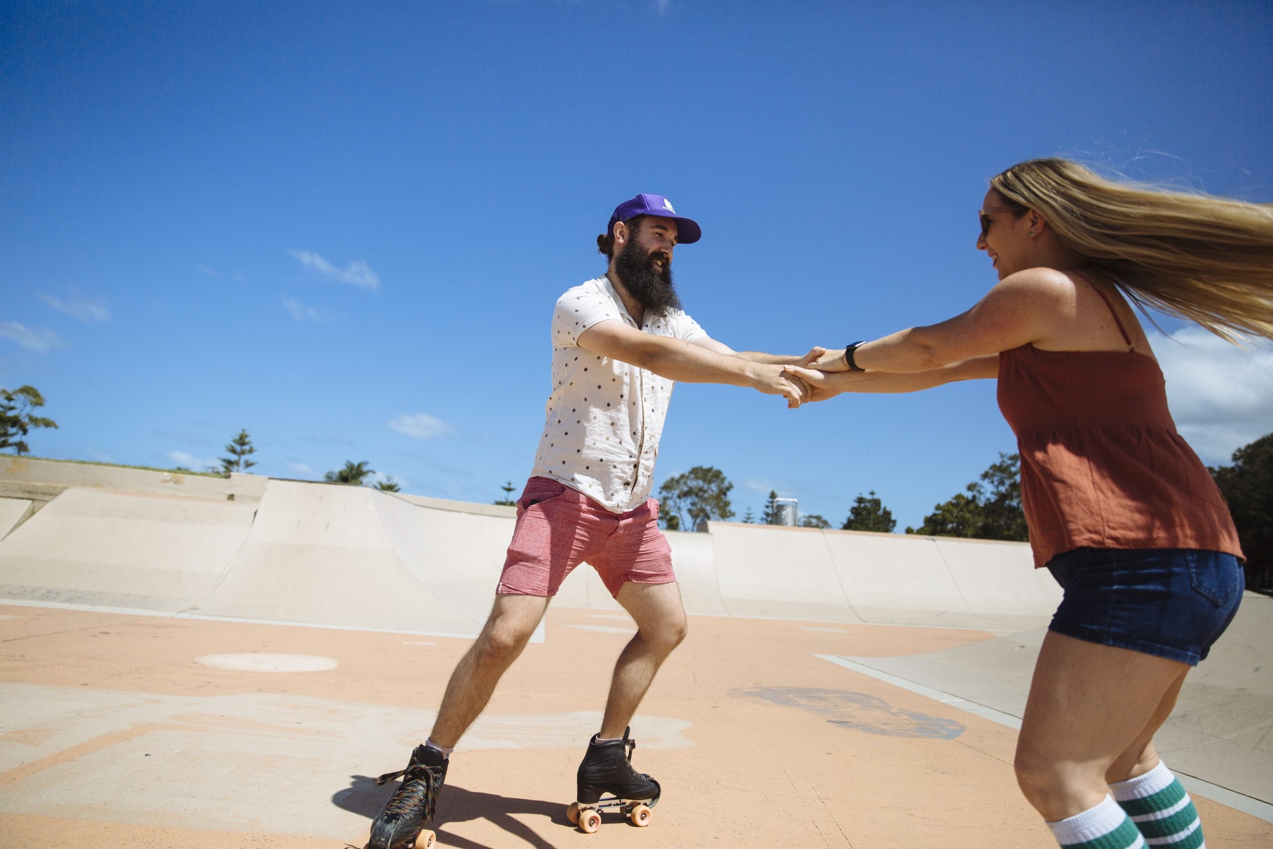 Matt and Tracey hold hands across from each other and are skating in circle at a skatepark with bright blue sky.